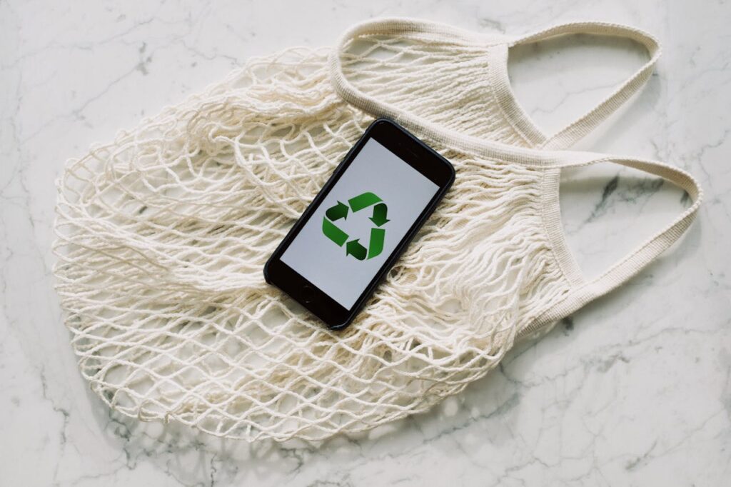 Top view of smartphone with recycling sign on screen placed on white textile mesh bag on white marble table in light room