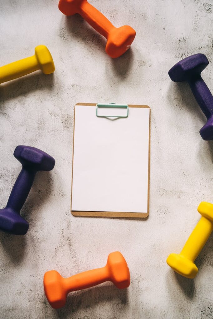 Top view of colorful dumbbells encircling a blank clipboard, offering copy space for text.